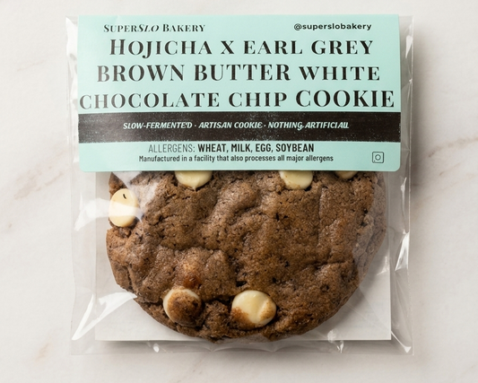 SuperSlo Bakery Hojicha Earl Grey Brown Butter White Chocolate Chip Cookie on a plate with tea leaves and a cup of tea.